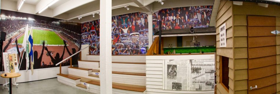 Puinen katsomo sisätilassa museosalissa. Taustaseinällä on kuva jalkapallostadionista. Sivussa on puinen lipunmyyntikioski.