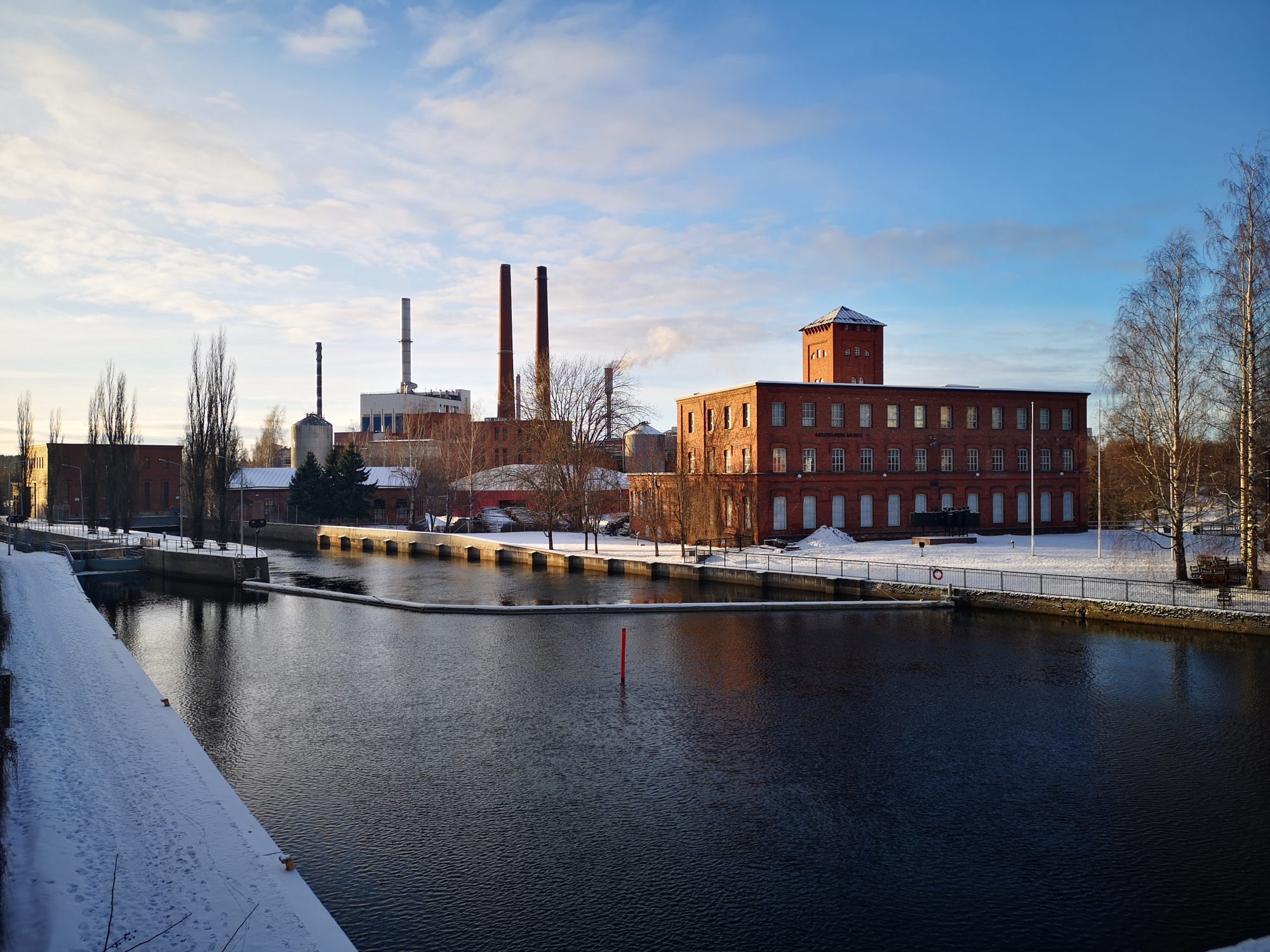 Kuvassa on talvi ja lunta maassa. Etulalla on järvi joka on sula. Taustalla tiilinen vanha rakennus sekä tehtaan piippuja.